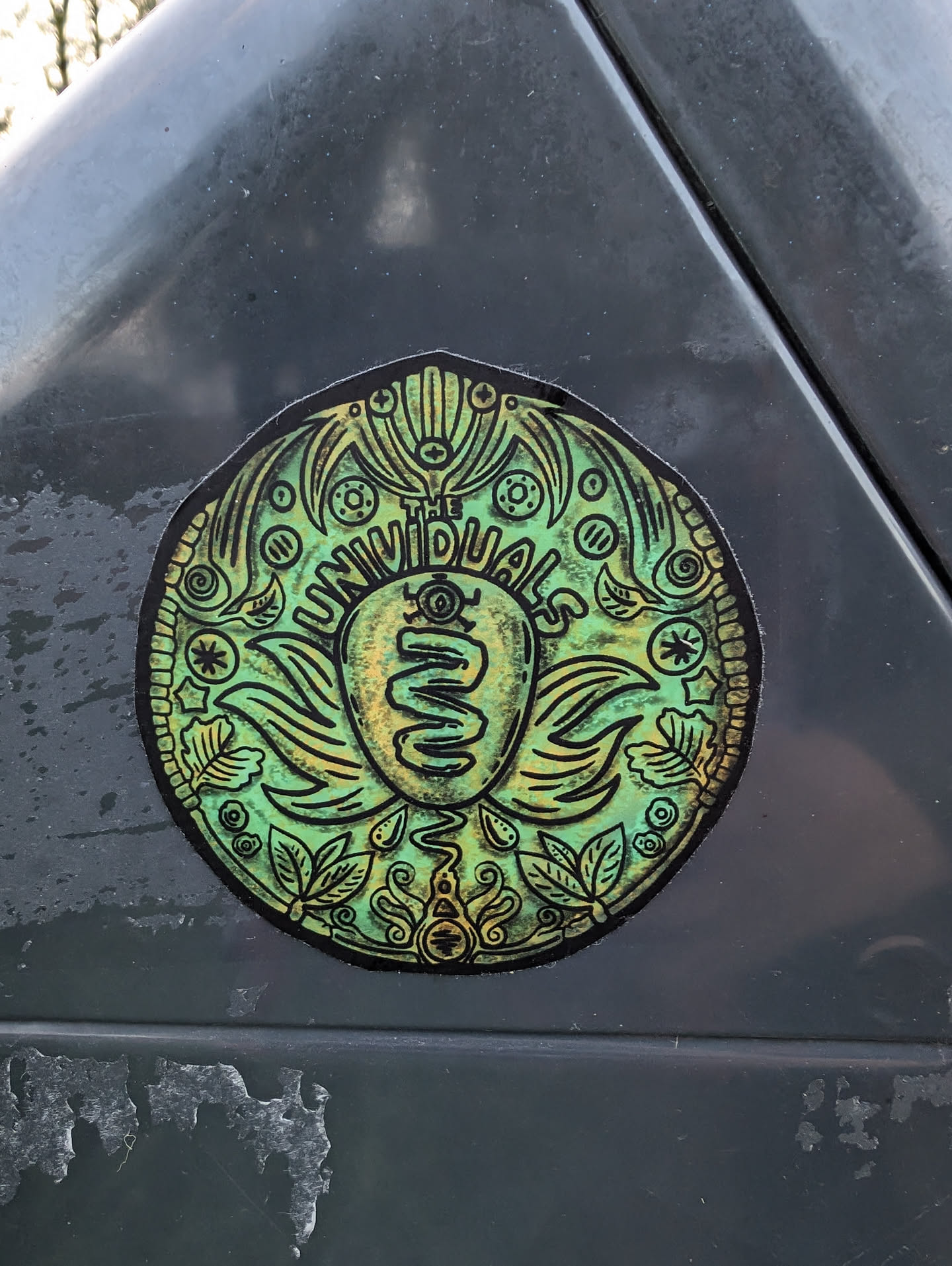A round sticker featuring a detailed design in shades of green and gold. The center depicts a stylized symbol, possibly a logo, surrounded by ornate botanical and geometric patterns. The text 'THE INDIVIDUALS' is visible within the design.