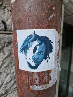 This sticker depicts a person with dark curly hair, their face partially obscured by a large, simple smiley face.  The colors are primarily shades of blue and white, creating a striking contrast. The sticker is affixed to a rusty metal pole, enhancing the urban aesthetic.