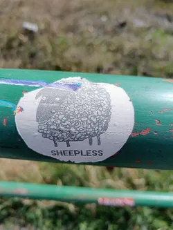 A sticker featuring a stylized sheep with the word 'SHEEPLESS' written below it. The sticker is placed on a weathered green metal surface. 