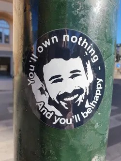 A round sticker featuring a stylized portrait of a smiling man with text that says "You'll own nothing and you'll be happy" around his face. The sticker is affixed to a dark green pole.