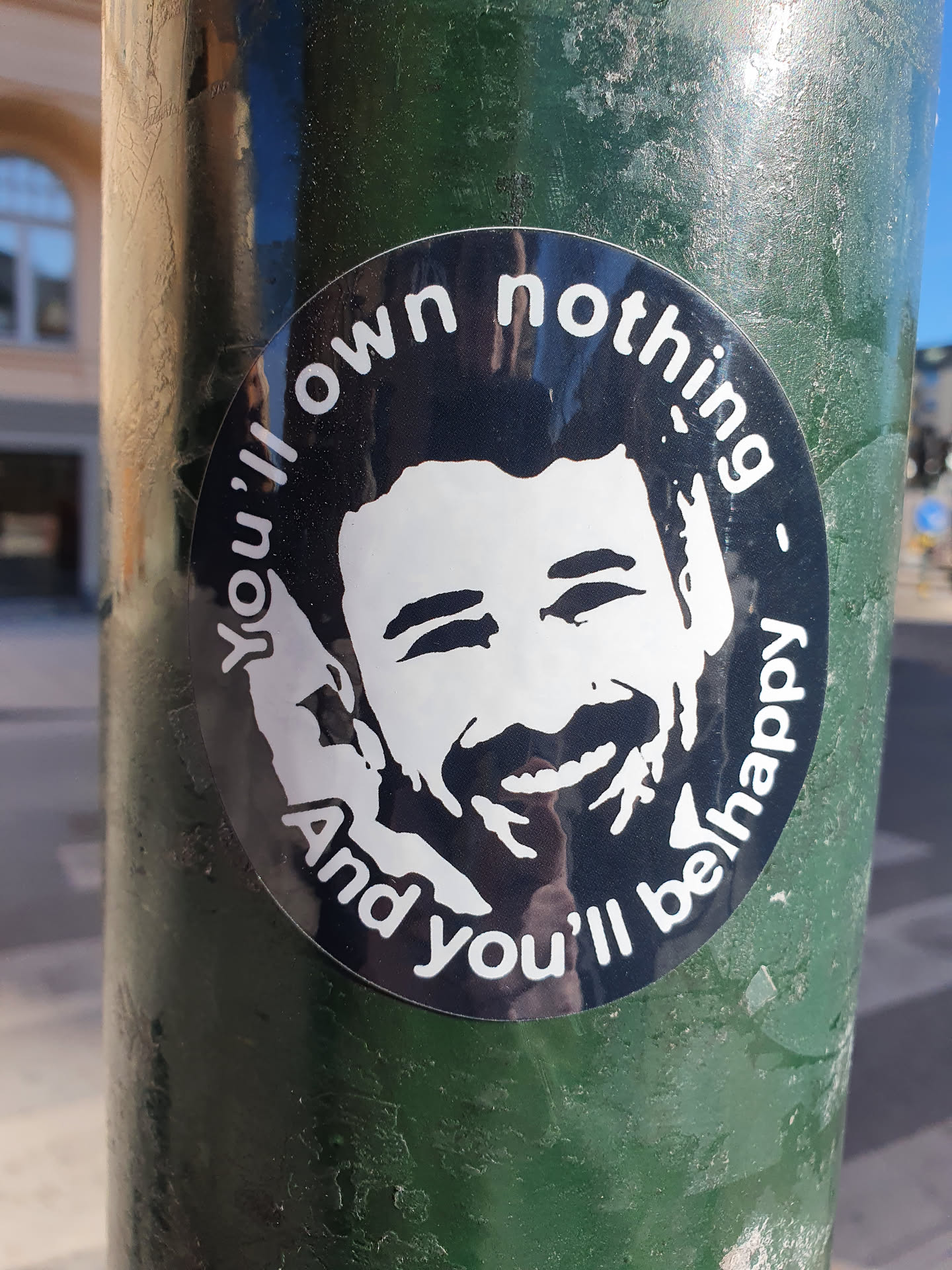 A round sticker featuring a stylized portrait of a smiling man with text that says "You'll own nothing and you'll be happy" around his face. The sticker is affixed to a dark green pole.