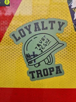 Green and black sticker with a military helmet, the words "LOYALTY" and "TROPA" and the phrase "TRUST JESUS" written on the helmet. 