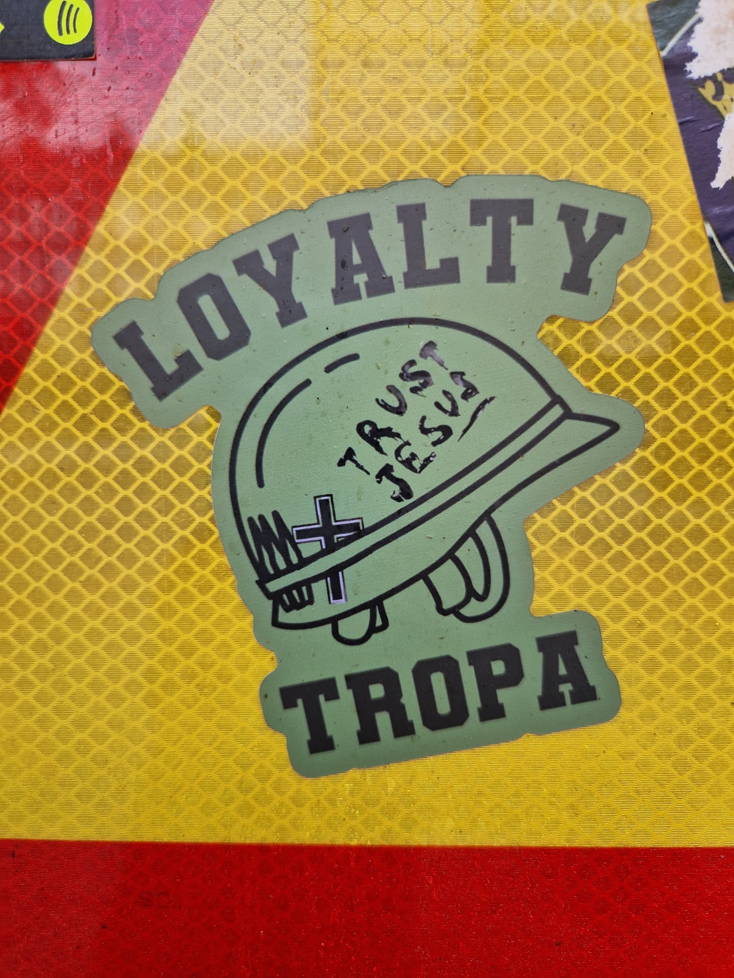 Green and black sticker with a military helmet, the words "LOYALTY" and "TROPA" and the phrase "TRUST JESUS" written on the helmet. 