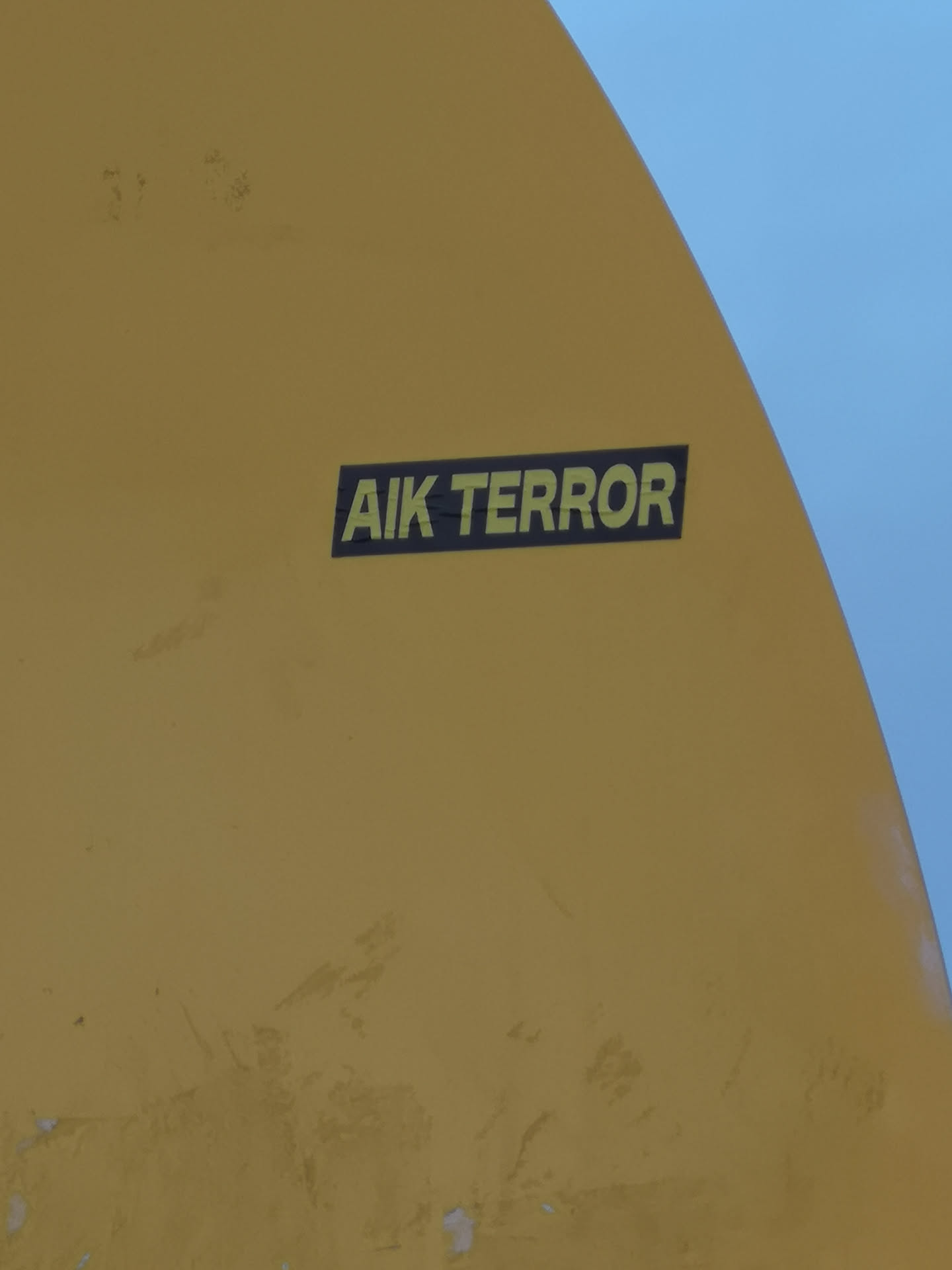 A rectangular sticker featuring the text 'AIK TERROR' in a bold, sans-serif, yellow-green font against a solid black background. The sticker is applied to a curved, mustard-yellow surface, possibly part of a vehicle or structure, with a clear blue sky visible in the upper right corner. The design is simple, high-contrast, and direct.