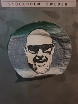 Round sticker featuring a stylized illustration of a bald man wearing sunglasses and a goatee. The artwork is in black and white, with a teal gradient background on the sticker.