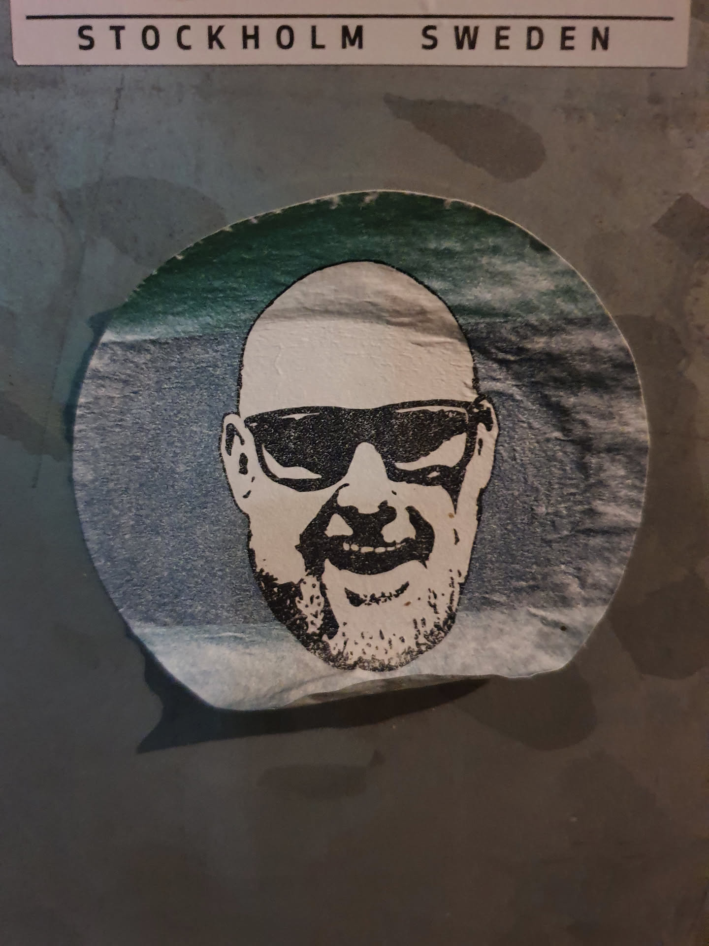 Round sticker featuring a stylized illustration of a bald man wearing sunglasses and a goatee. The artwork is in black and white, with a teal gradient background on the sticker.