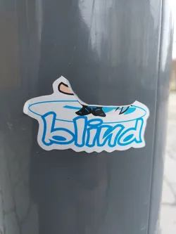 A sticker featuring the Blind skateboard brand logo. The logo is blue text on a white background. There is a small cartoonish illustration of legs and feet in the top of the sticker. The sticker is slightly damaged, and has been placed on a gray pole.