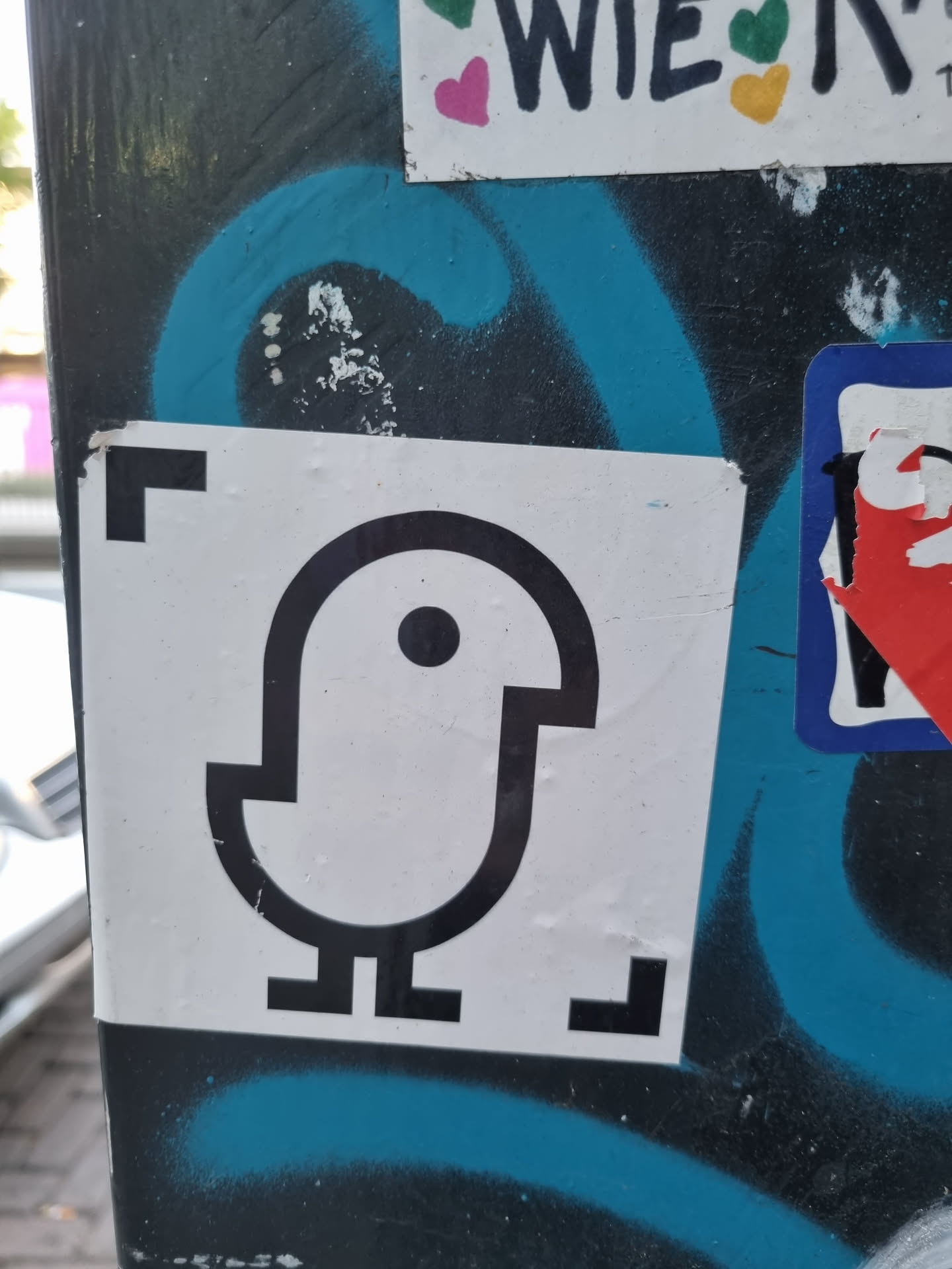 A simple, black and white sticker featuring an abstract bird illustration. The sticker has a minimalist design with clean lines and a single spot for an eye. It is stuck on a surface with other stickers and graffiti.