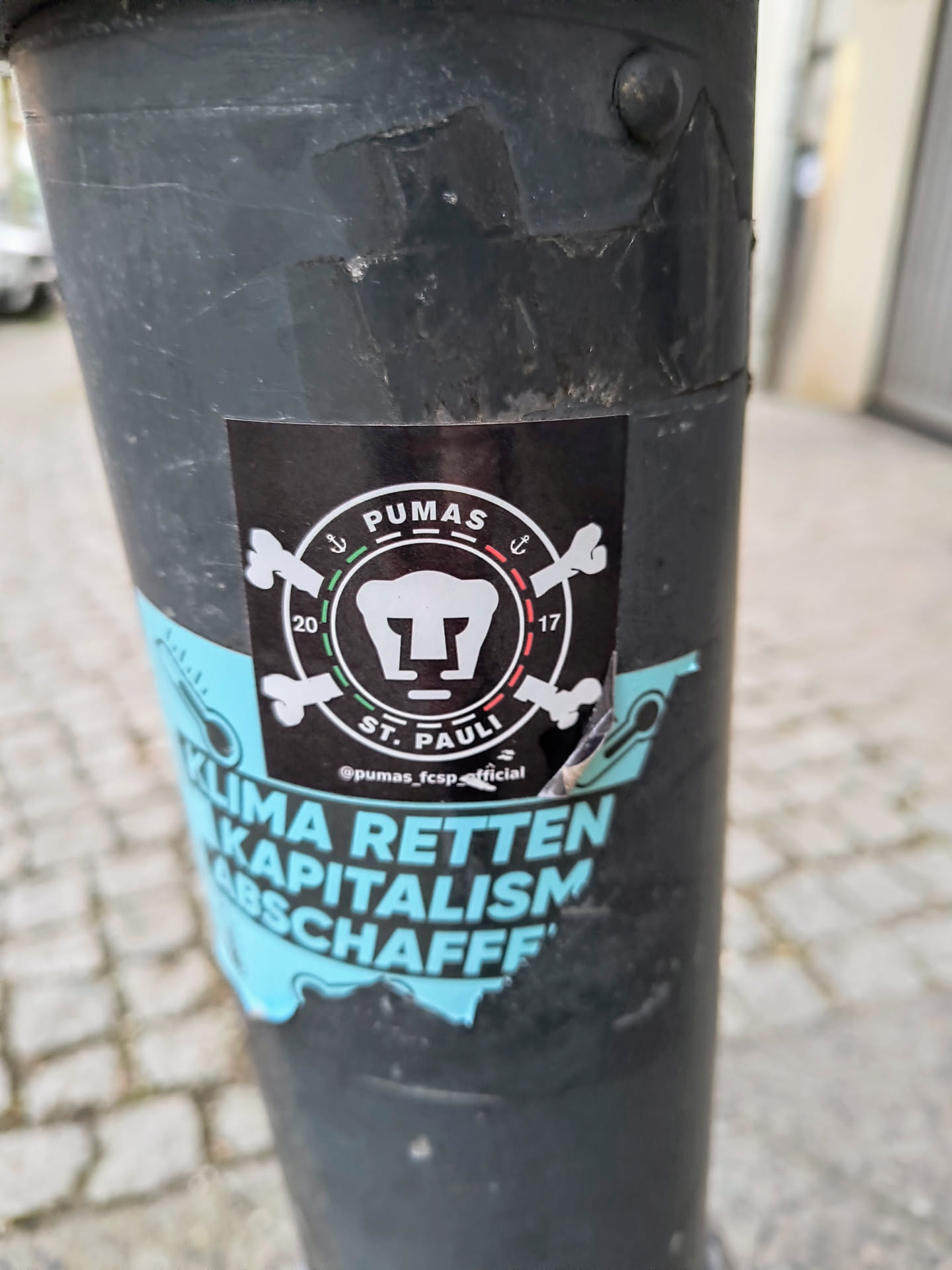 This is a black sticker with the words PUMAS ST. PAULI in white text. The sticker also features an image of a stylized anchor and bones, along with the years 2017 and 2020. The sticker is located on a pole.