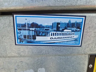 A sticker featuring a monochrome image of Djurgårdens IF boat in Stockholm. The boat is prominently displayed against the backdrop of the city's skyline. The sticker is rectangular with a blue border.