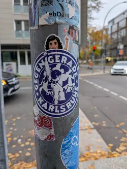 A circular sticker primarily in navy blue and white, featuring a custom die-cut top showing an illustrated girl with wavy dark hair raising her fist. Within the round seal, a silhouette of a supporter is raising their arm against a dynamic background. Encircling this graphic is a navy border with white text reading 'GEGENGERADE' on top and 'KARLSRUHE' on the bottom, flanked by small five-pointed stars.