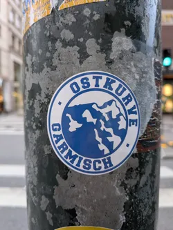 Street sticker A circular sticker adhered to a weathered, dark metal pole. The sticker features a blue border outlining a white circular band with the uppercase blue text 'OSTKURVE' arched over the top and 'GARMISCH' arched along the bottom, separated by two small blue square dots. In the center, a blue circle displays a stylized graphic of snowy, jagged mountain peaks.