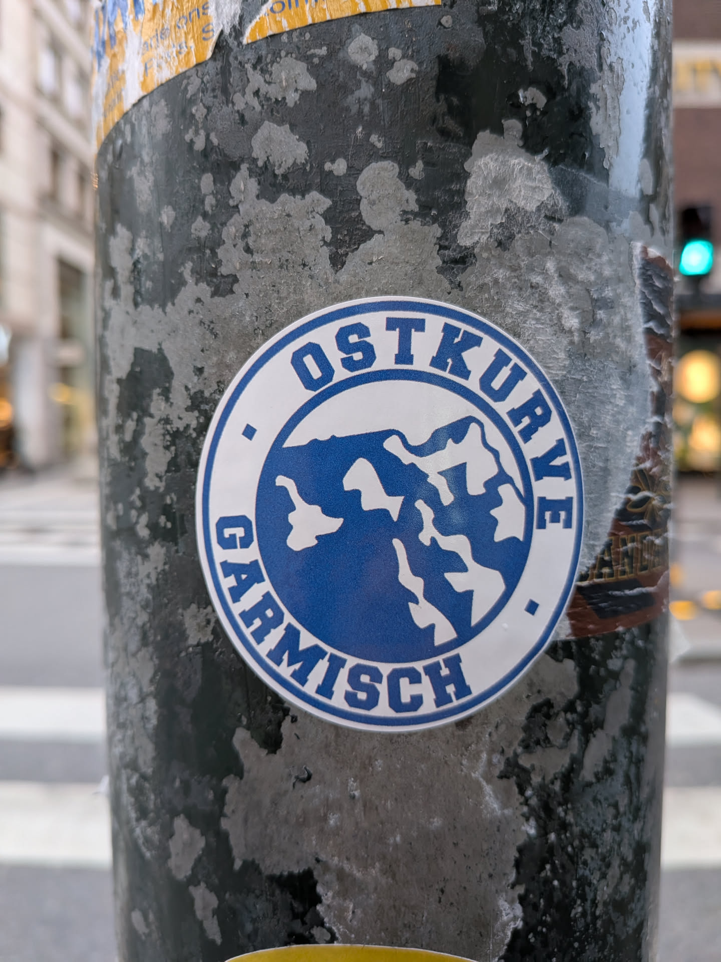 A circular sticker adhered to a weathered, dark metal pole. The sticker features a blue border outlining a white circular band with the uppercase blue text 'OSTKURVE' arched over the top and 'GARMISCH' arched along the bottom, separated by two small blue square dots. In the center, a blue circle displays a stylized graphic of snowy, jagged mountain peaks.