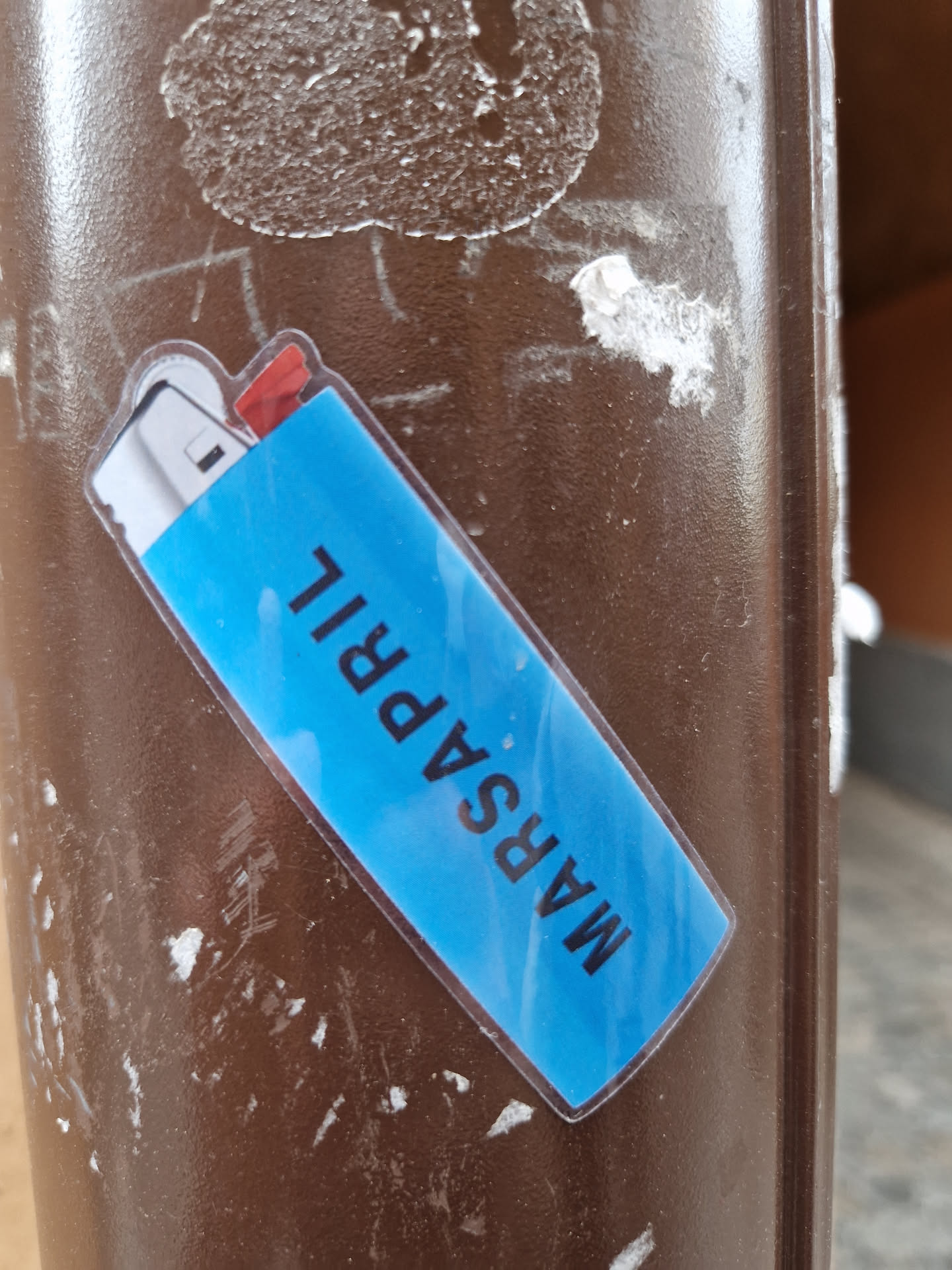 A blue sticker shaped like a lighter with the text "MARSAPRIL" written vertically on it.  The sticker is attached to a brown surface.