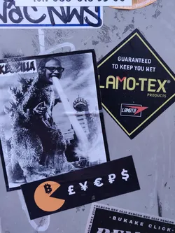 A sticker featuring a black and white image of Godzilla seemingly breathing fire onto a person wearing glasses.  Below is a smaller sticker with a Pac-Man-like shape, containing a Bitcoin symbol and various currency symbols.