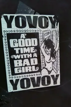 A rectangular sticker featuring a black and white design. The sticker has a cracked and aged appearance, suggesting it has been in place for some time. The design includes a drawing of a woman, and bold lettering that reads "A GOOD TIME WITH A BAD GIRL". The top and bottom of the design are framed by the word "Yovoy".