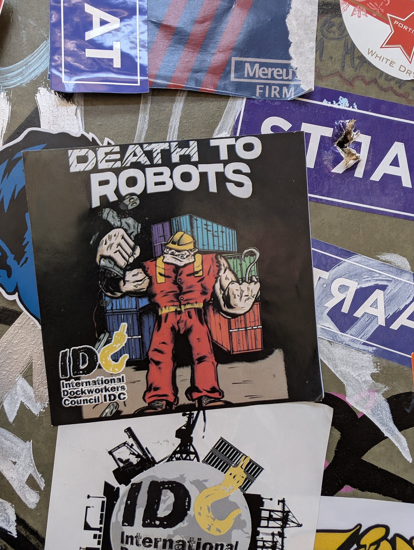 A sticker featuring a muscular worker surrounded by shipping containers, with the text "DEATH TO ROBOTS" above and "International Dockworkers Council IDC" below. The style is reminiscent of protest art or union imagery.